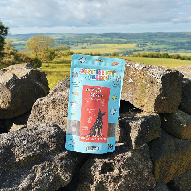 Front view of a packet/ pouch of 'Just'ere for treats' natural beef jerky pack of 4 on a old stone wall with an english countryside background